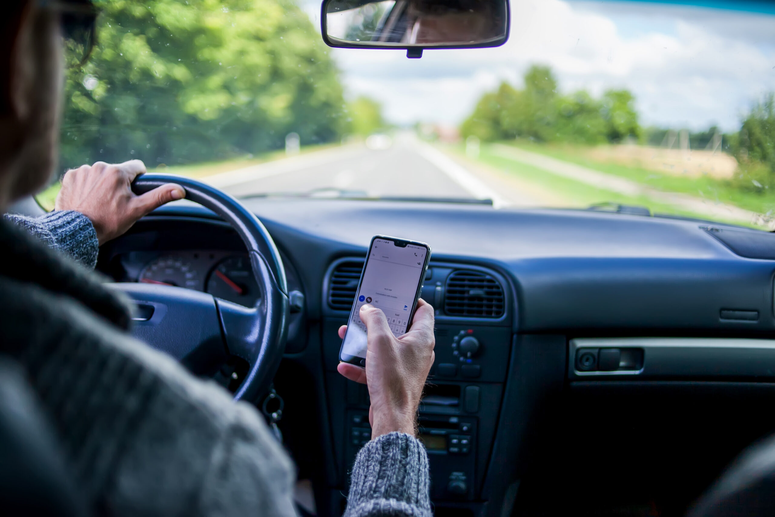 Driver holding a smartphone while driving on a road, illustrating the dangers of distracted driving for work.