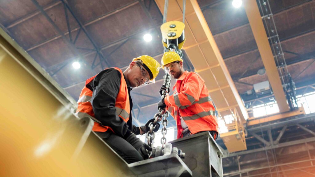 Two male industrial workers wearing safety helmets and high-visibility vests operate heavy lifting equipment in a factory, representing the connection between men’s health and workplace safety during Movember.