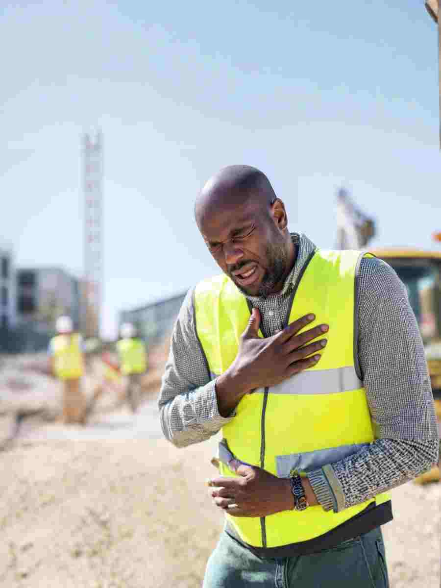 A construction worker wearing a high-visibility vest clutching his chest on-site, illustrating the importance of recognising cardiovascular health risks in the workplace.