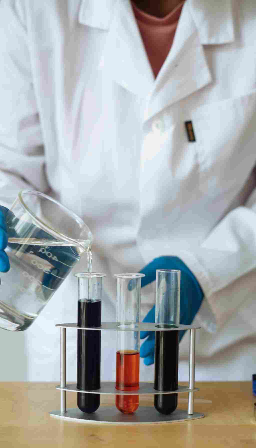 Laboratory Chemical Testing and Analysis Procedures Lab technician in a white coat and gloves pouring liquid from a beaker into test tubes containing different coloured chemical samples.