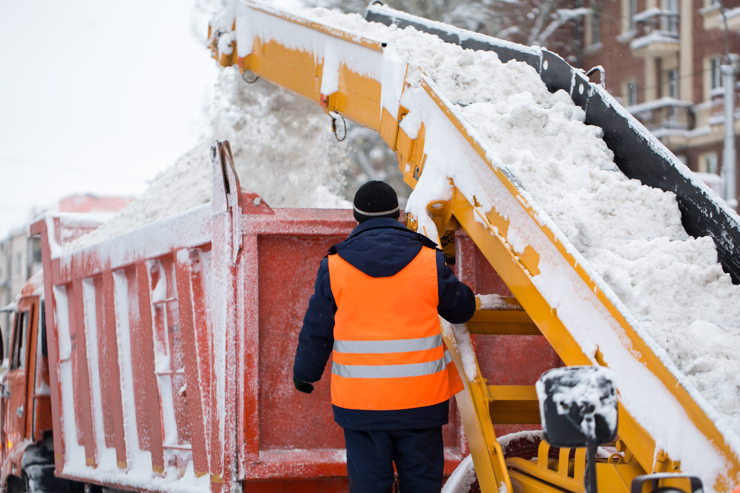 Winter Roadwork Safety: Snow Removal Operations Worker in a high-visibility vest operating snow-removal machinery and loading snow into a truck during winter conditions.