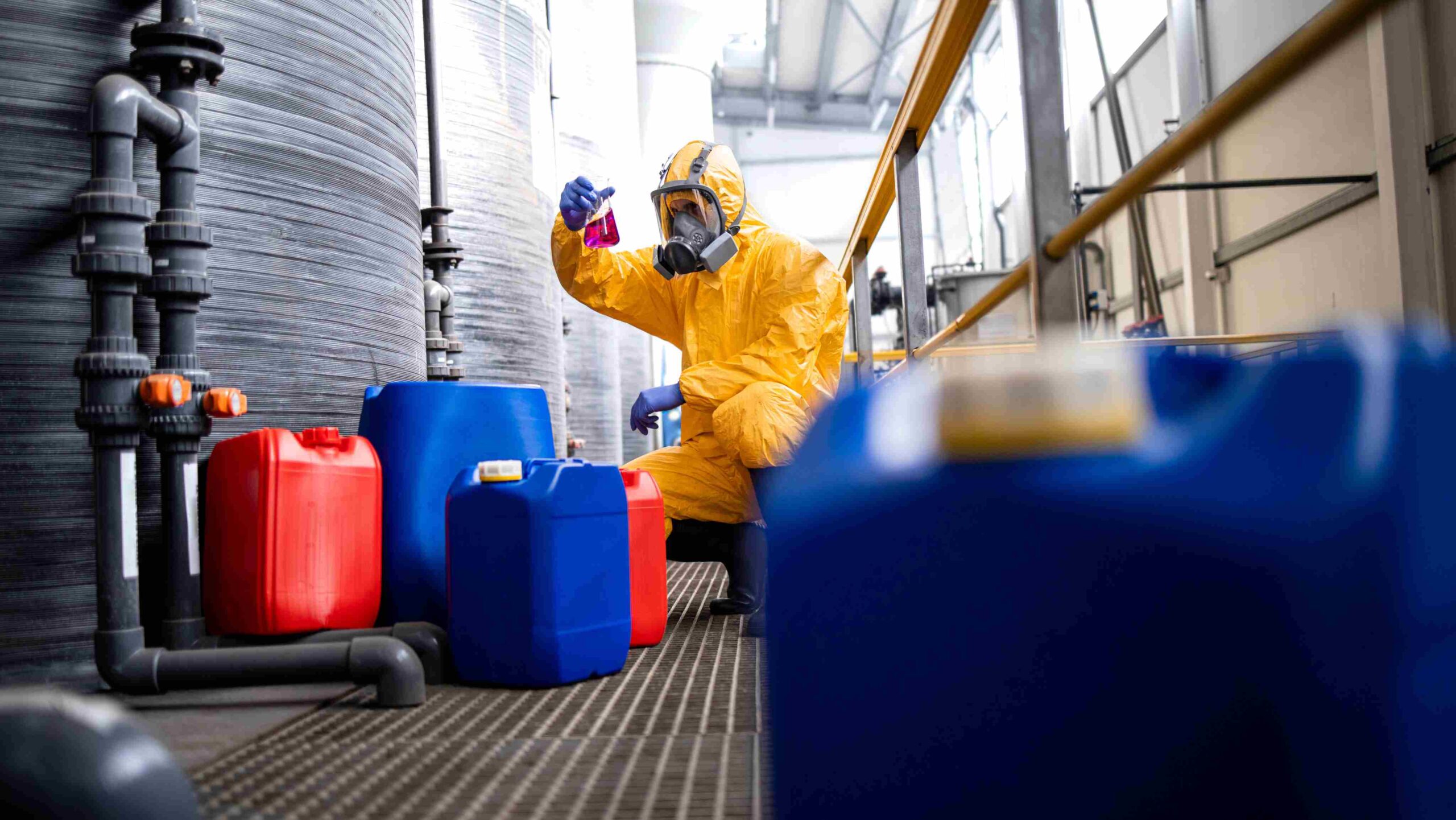 Chemical Sampling and Hazardous Substance Inspection in Industrial Facility Worker in a yellow hazmat suit examining a sample of coloured liquid near red and blue chemical containers in an industrial setting.