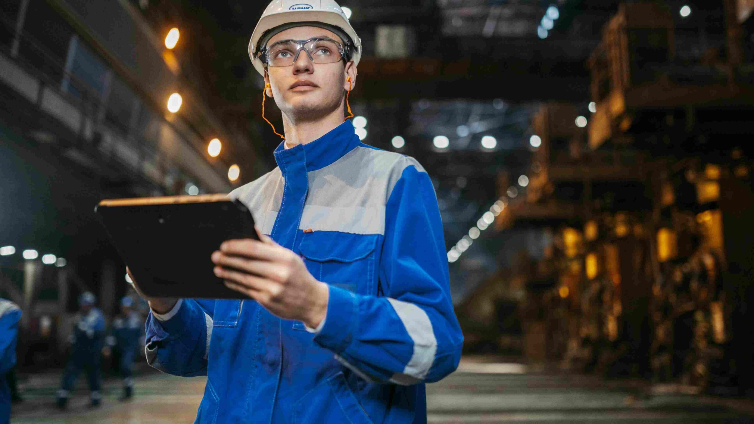 A worker in a hard hat and protective eyewear holding a tablet while conducting a safety inspection inside an industrial facility.