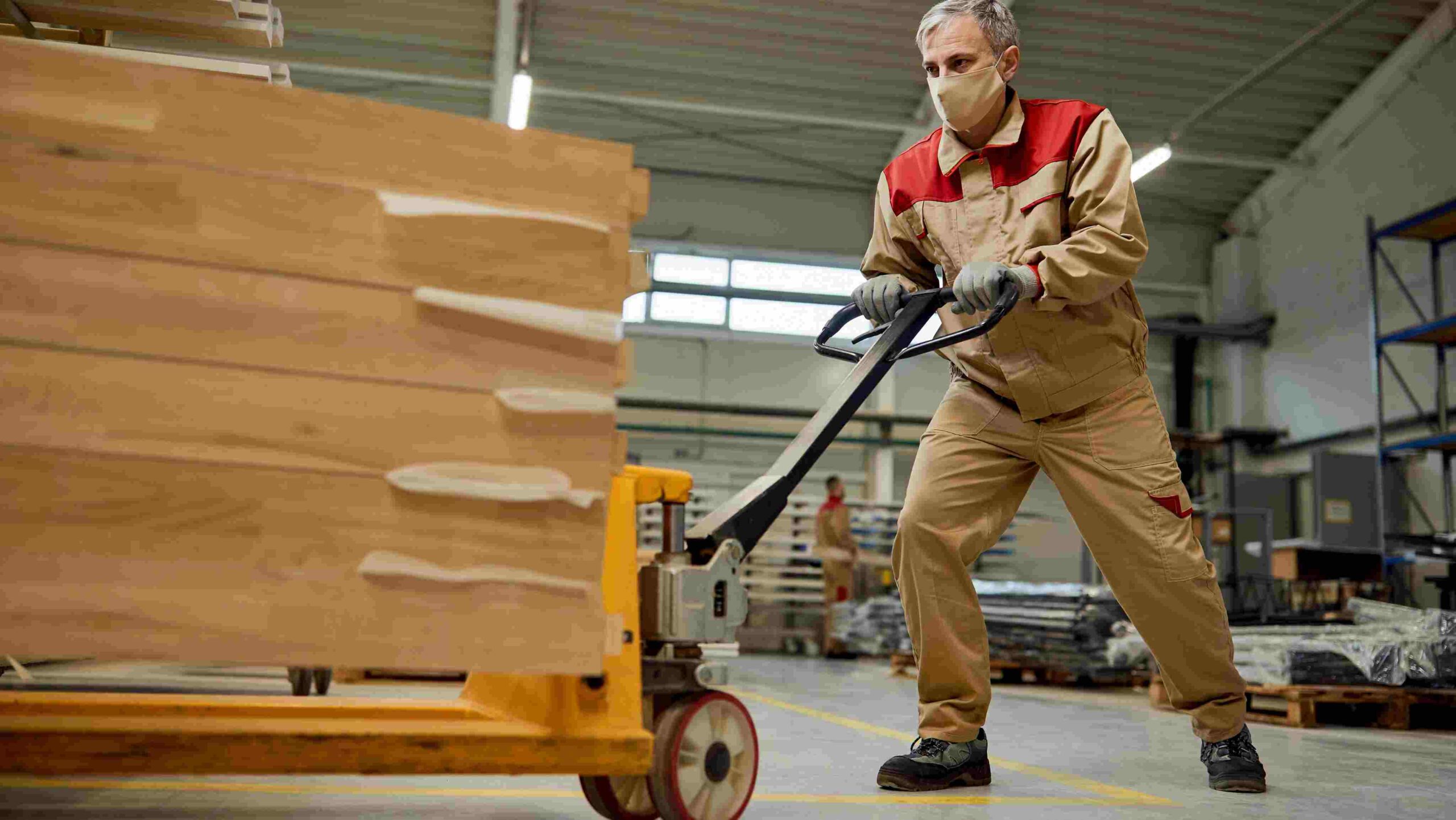 Worker using a pallet jack to move a heavy load of timber in a warehouse, demonstrating safe mechanical handling to reduce manual lifting risks.