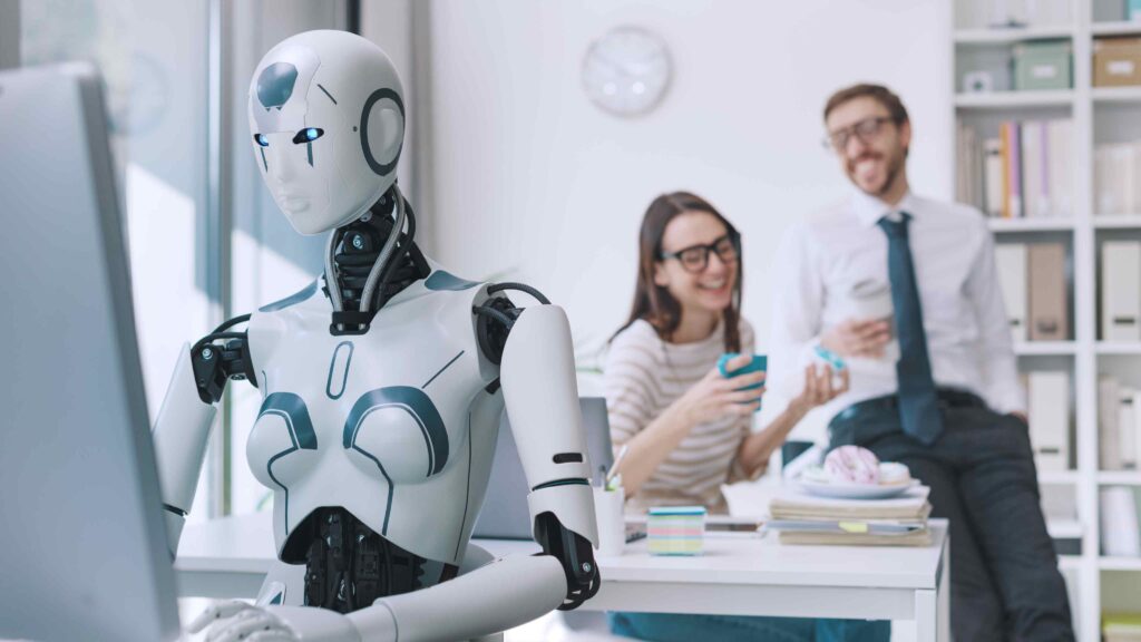Humanoid robot working at a computer in a modern office while two employees interact in the background, representing AI and robotics in workplace safety.