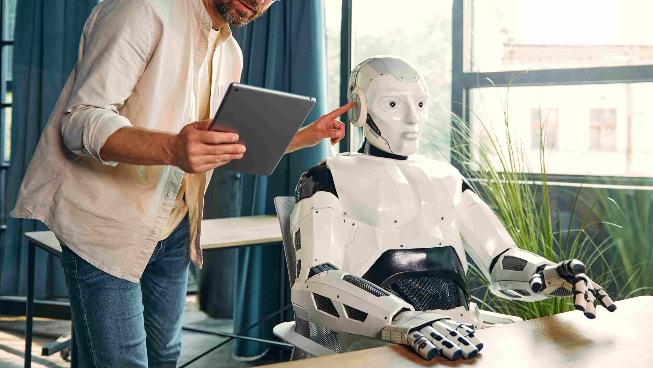 Training and Integrating Robotics in the Modern Workplace Man using a tablet while training or configuring a humanoid robot seated at a desk in a modern office.