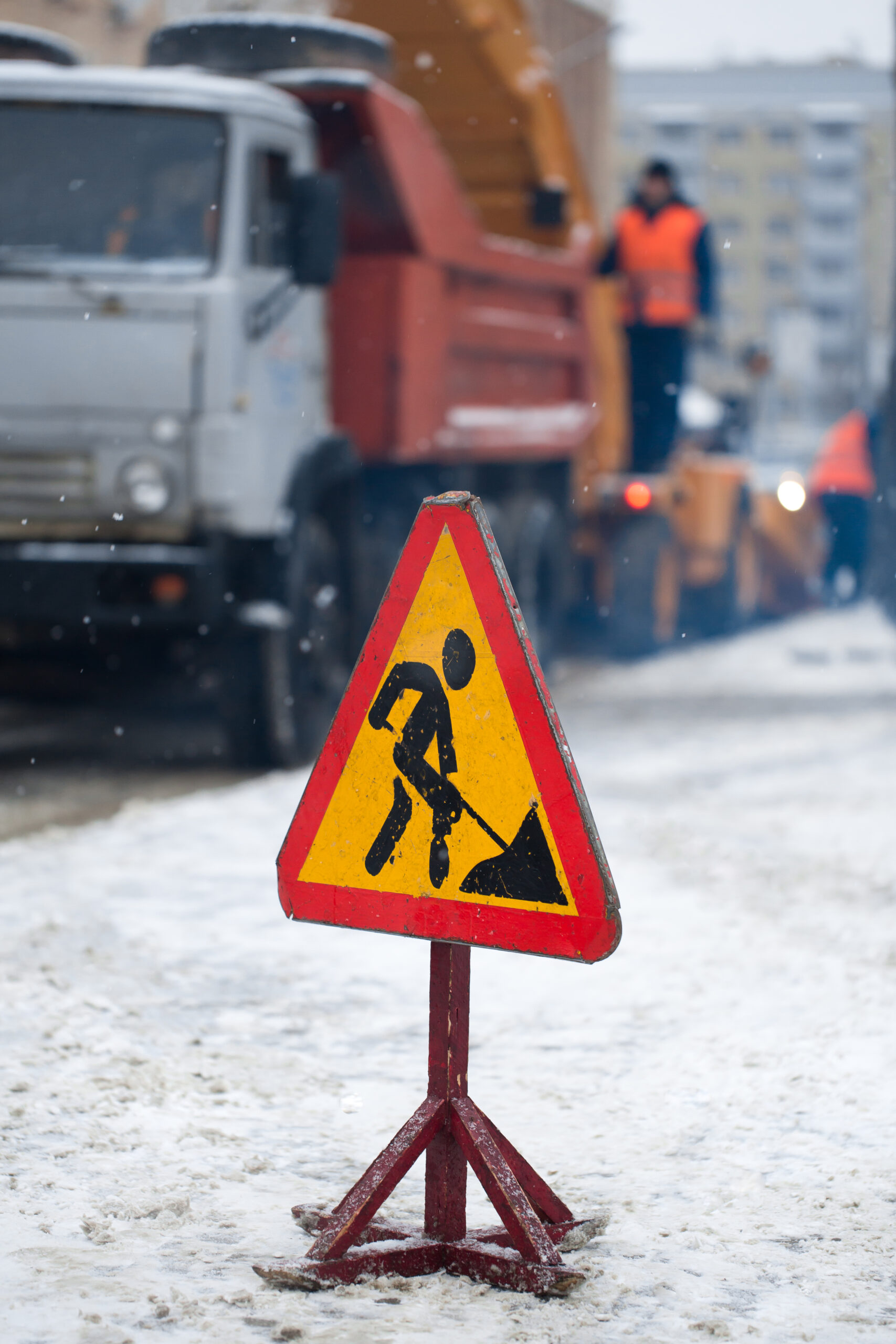 Winter Roadwork Safety Sign in Snowy Conditions Roadwork warning sign on a snowy street with workers and machinery operating in the background.