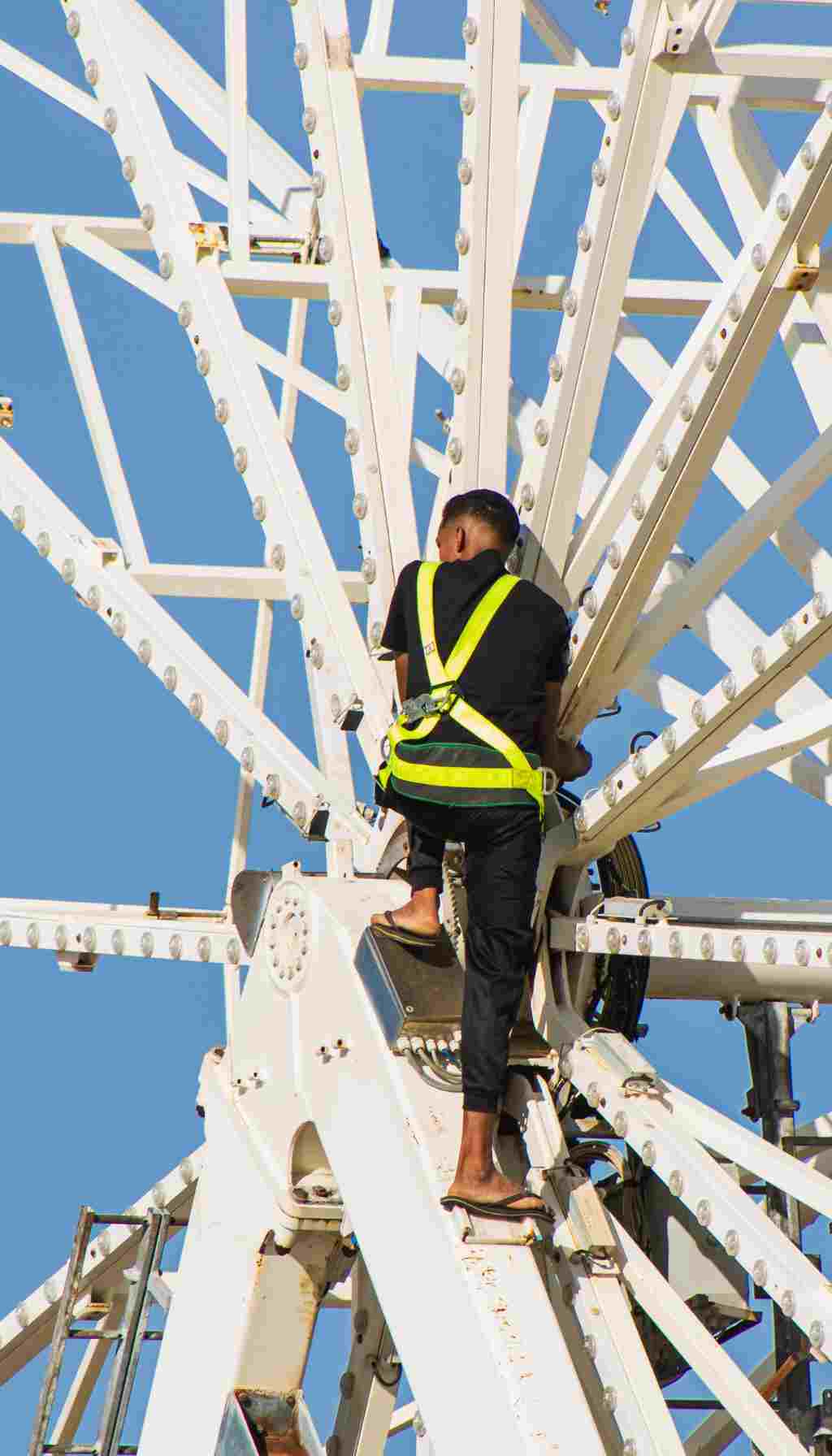 Worker on a high metal structure wearing a safety harness while performing maintenance at height under clear sky conditions.