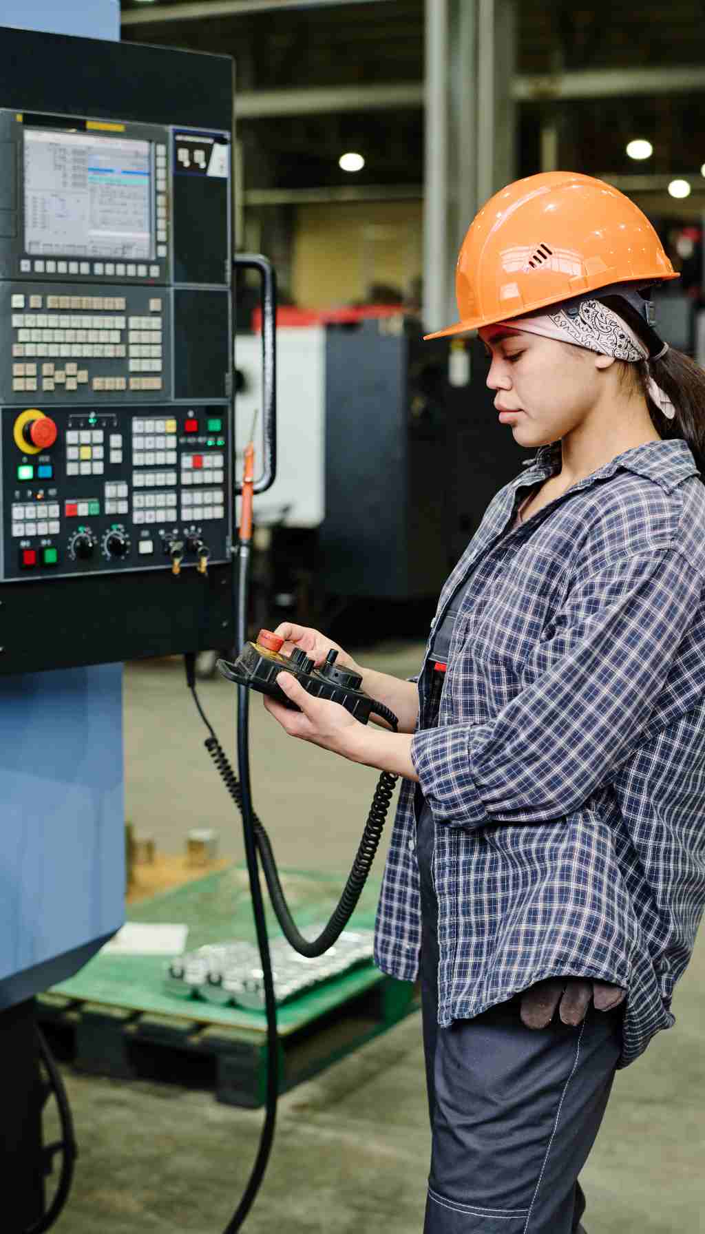 A worker wearing a hard hat using a handheld control device to operate CNC machinery inside an industrial manufacturing environment.