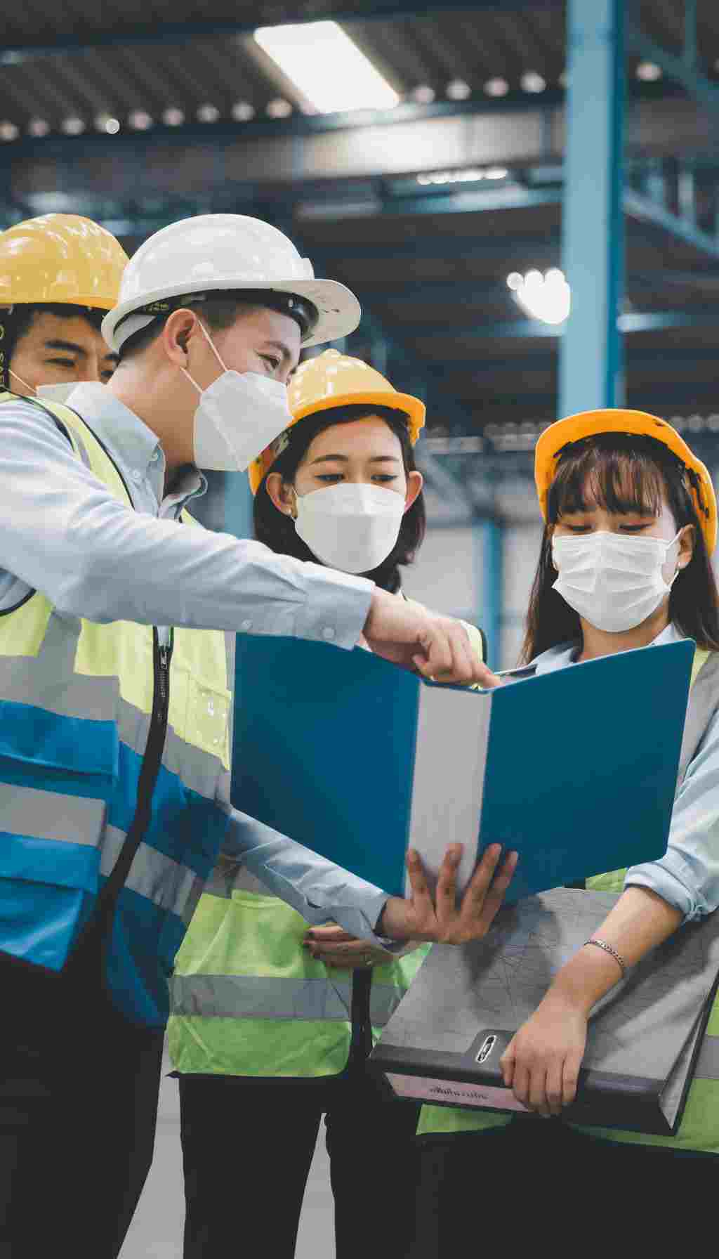 Team Reviewing Safety Procedures on the Worksite Workers wearing hard hats, safety vests, and face masks reviewing safety documents together inside an industrial facility.