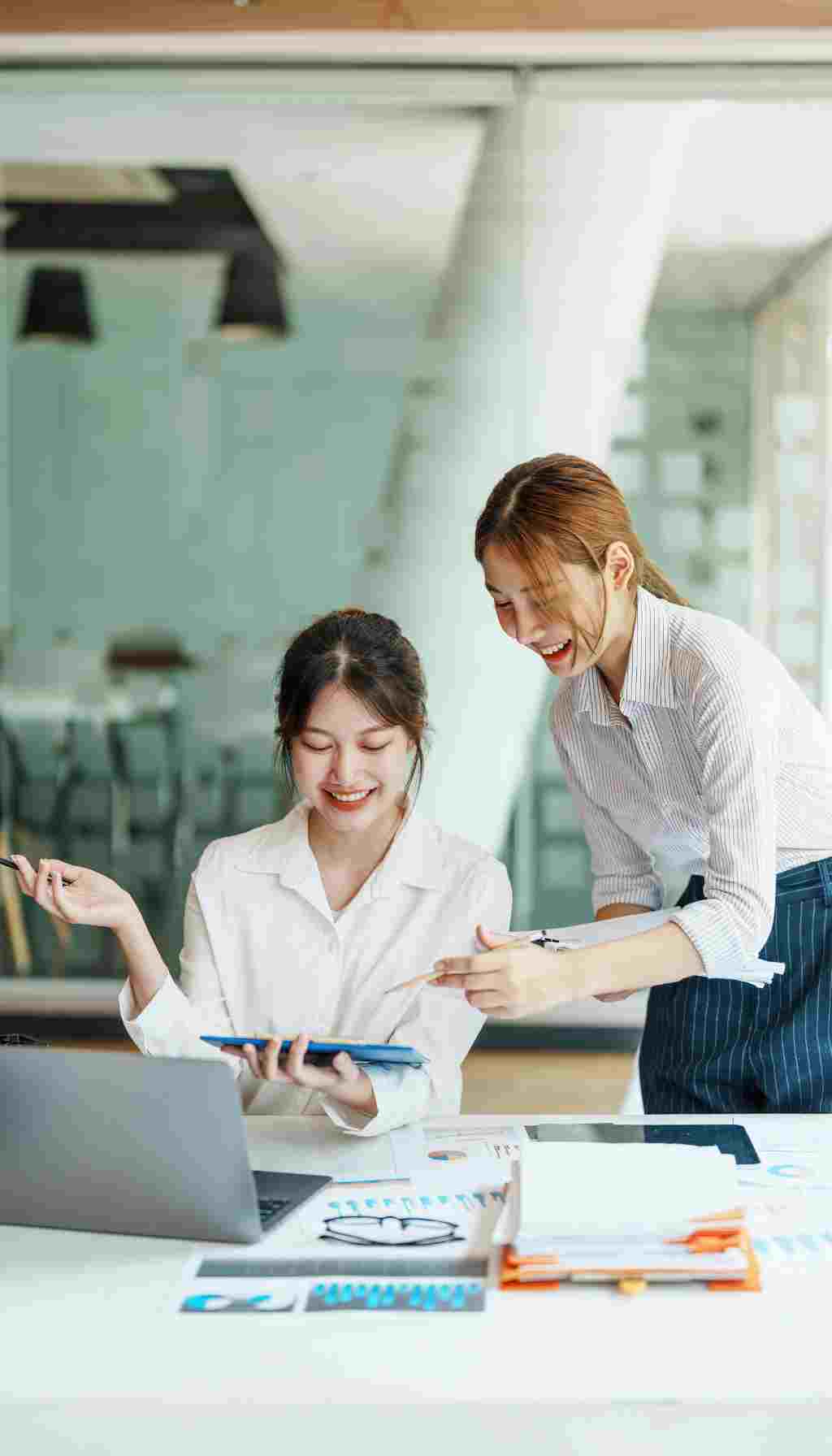 Two colleagues collaborating at a desk with documents, charts, and a laptop, discussing workplace planning and safety processes.