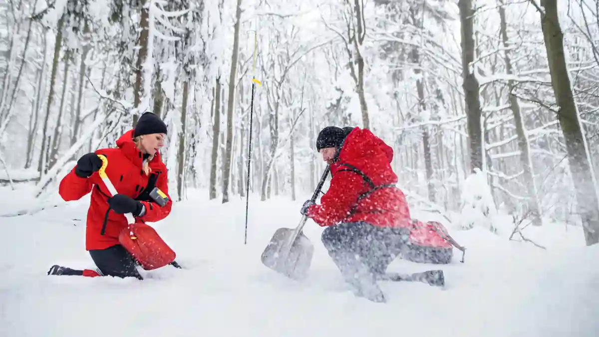 Two rescue workers in red jackets digging in deep snow during a winter emergency operation in a forest.