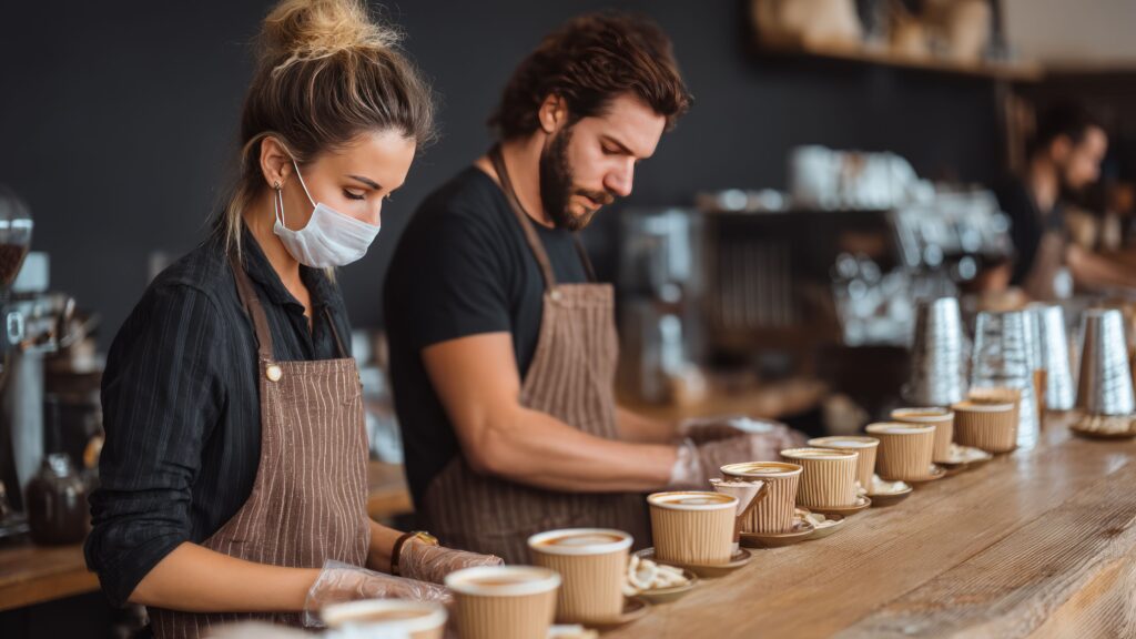 Two bartenders wearing aprons prepare multiple coffee drinks in a busy bar; one bartender wears a face mask and gloves, highlighting food safety and allergen handling.