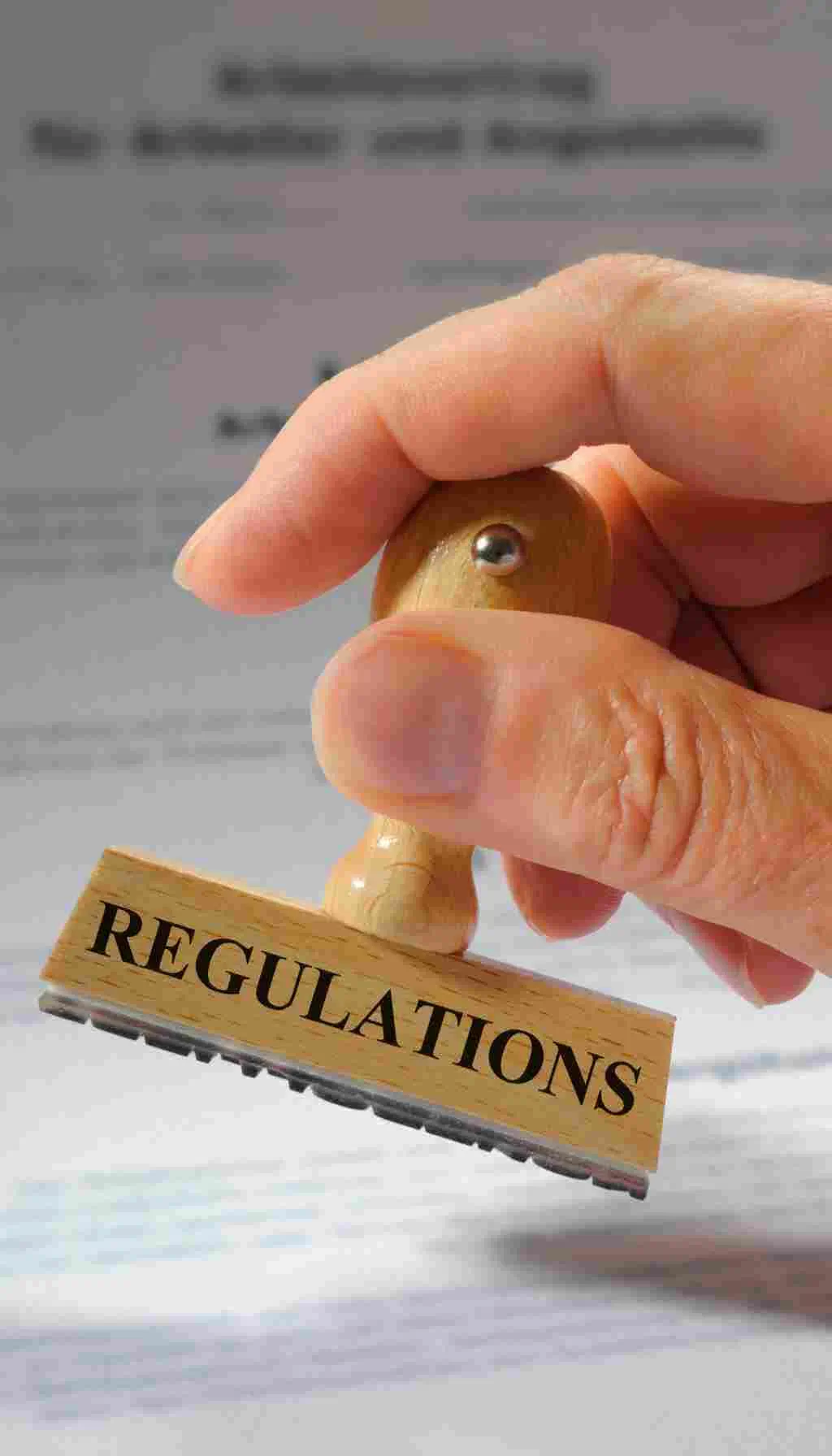 Hand holding a wooden stamp marked “Regulations” over official compliance documents.