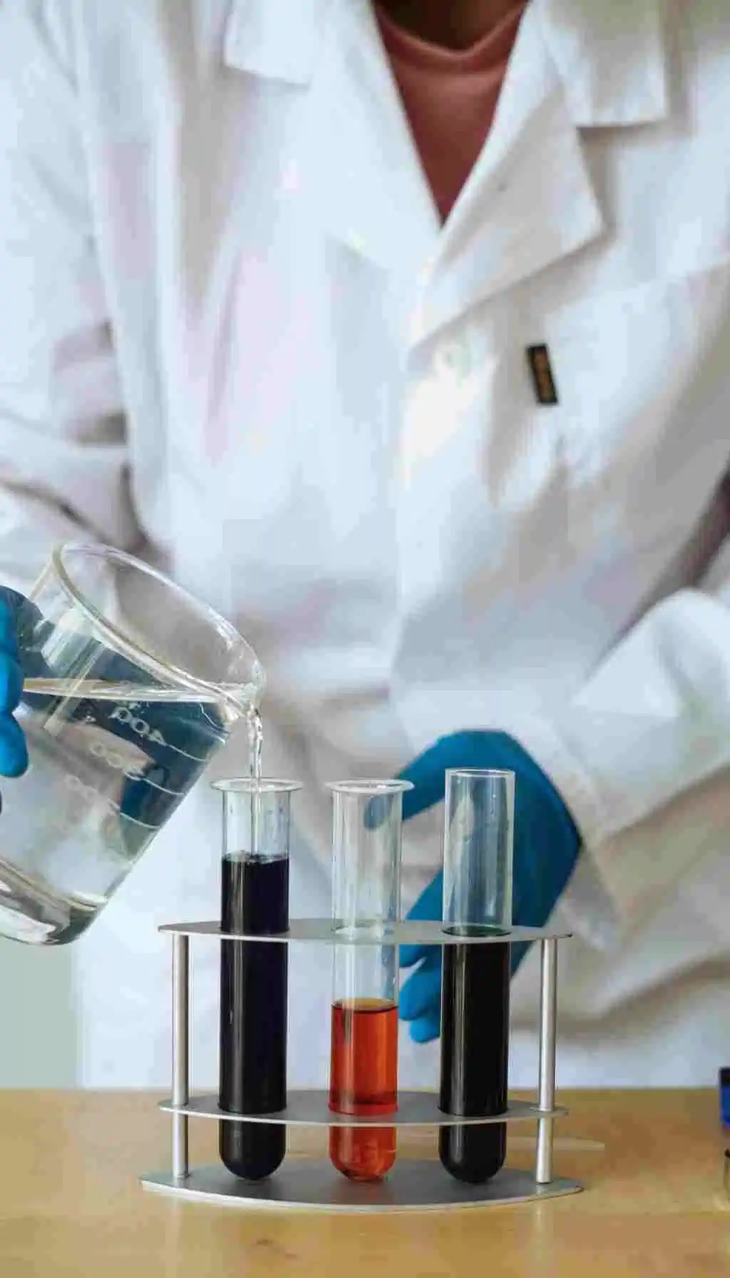 Lab technician in a white coat and gloves pouring liquid from a beaker into test tubes containing different coloured chemical samples.