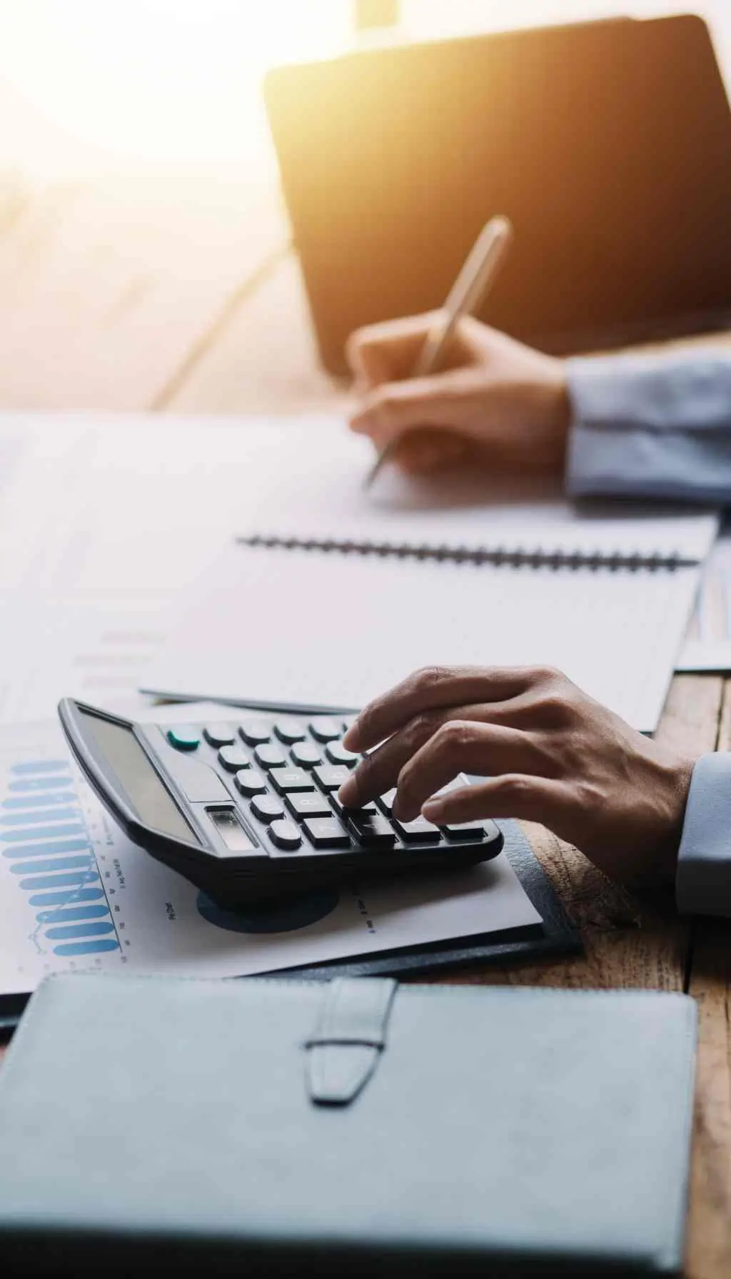 Business Professional Calculating Finances with Calculator and Reports Hands using a calculator and writing on financial documents at a desk, with charts and reports visible.