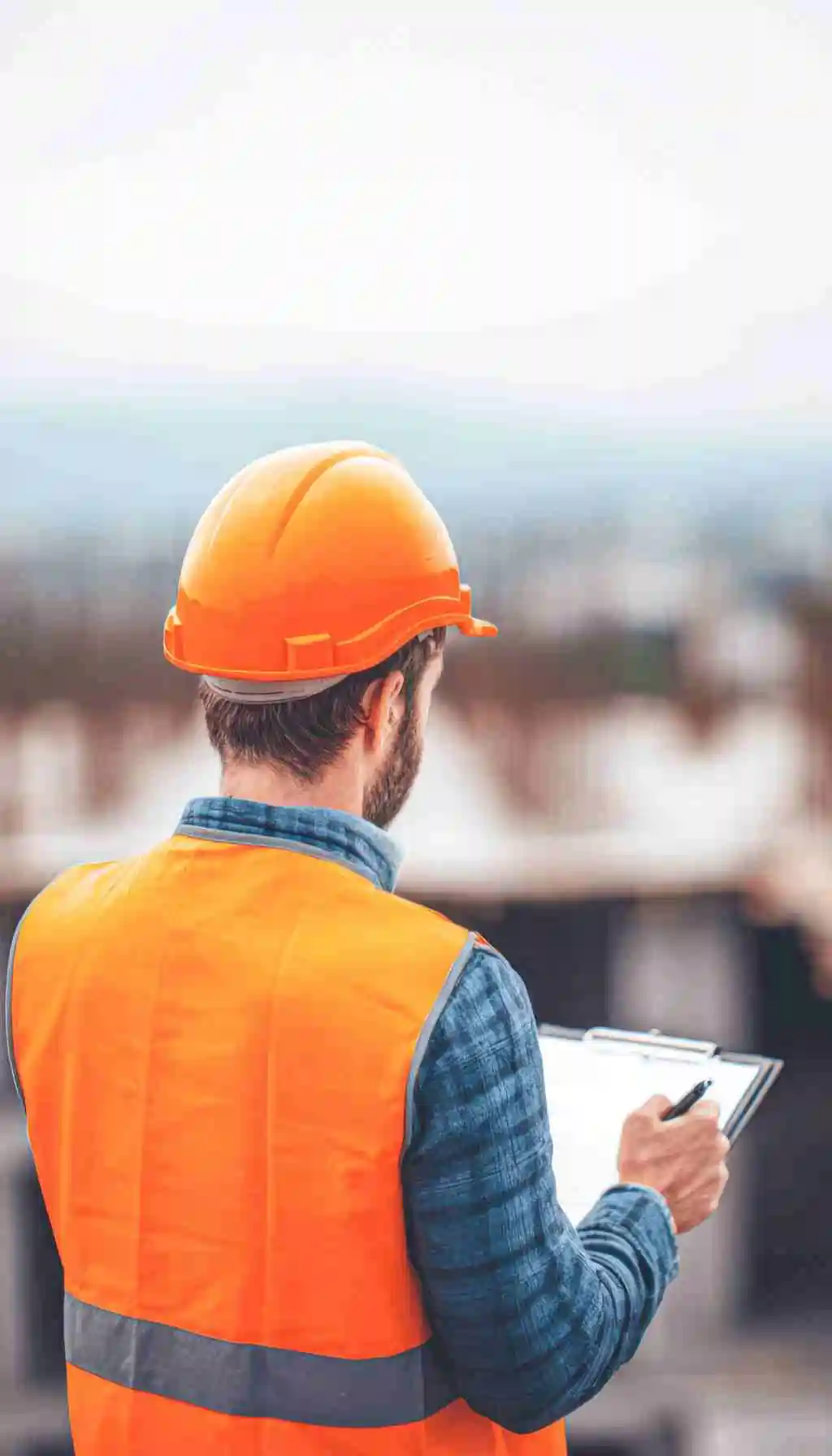 Construction site inspector wearing orange safety vest and hard hat reviewing checklist during site inspection.