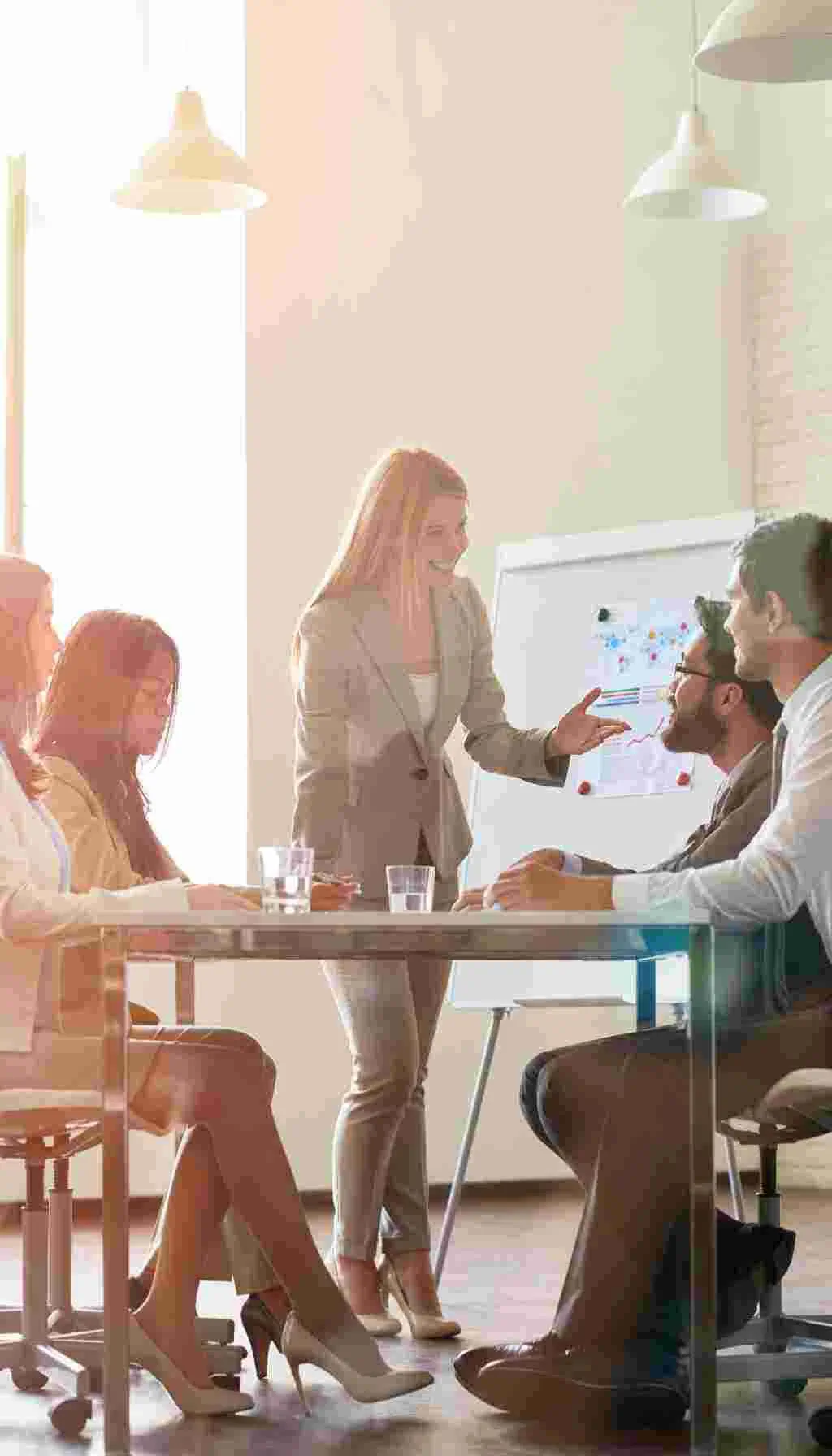 A team of professionals in a bright office discussing quality management strategies during a leadership meeting, with a presenter standing beside a flip chart.