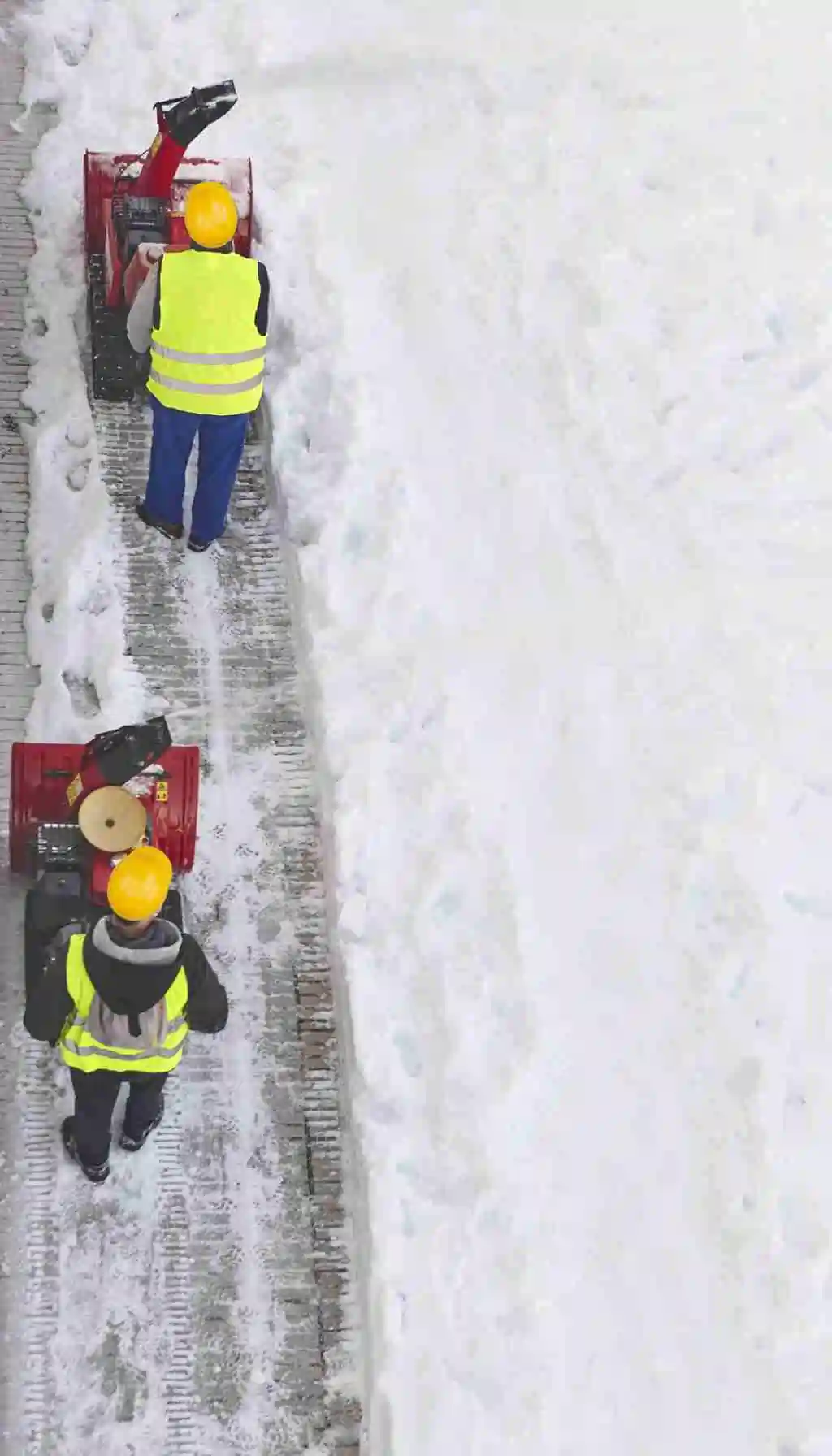 Two workers wearing high-visibility vests and helmets operating snow blowers to clear a snowy footpath.