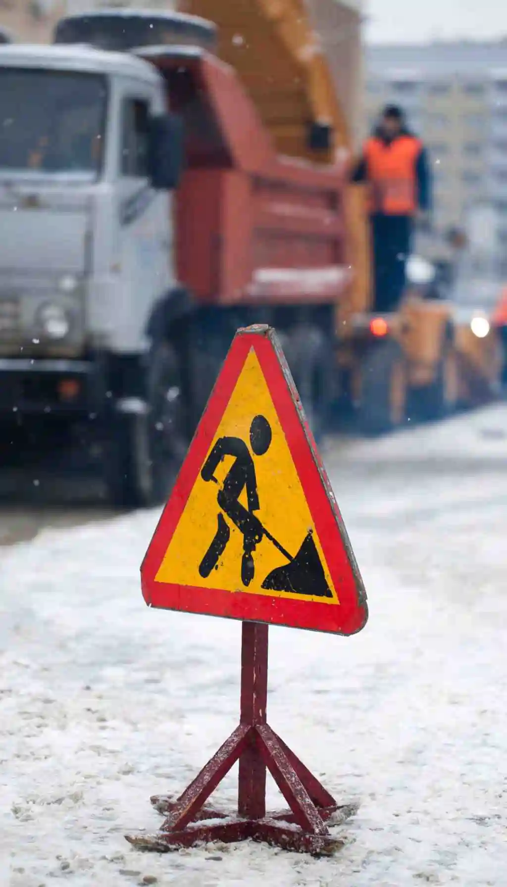 Roadwork warning sign on a snowy street with workers and machinery operating in the background.