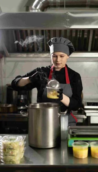 Chef wearing gloves and a uniform ladling hot soup with vegetables into takeaway containers in a commercial kitchen, demonstrating safe portioning and hot-holding practices.