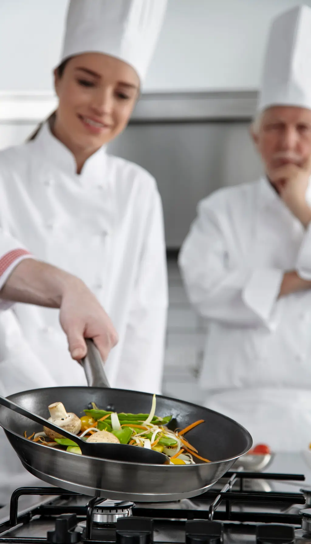 Chef cooking vegetables in a frying pan while a senior chef supervises in a professional kitchen.