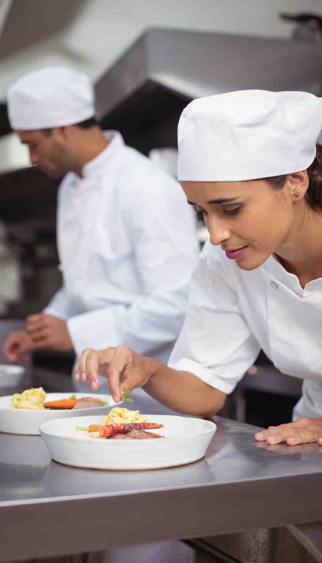 Two chefs in white uniforms carefully plating dishes in a commercial kitchen, demonstrating professional food preparation and hygiene standards.