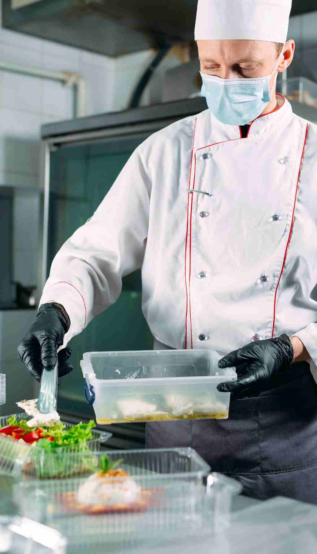 Chef Safely Packaging Prepared Meals in a Commercial Kitchen Chef wearing a mask and black gloves placing cooked food into plastic meal containers in a commercial kitchen.