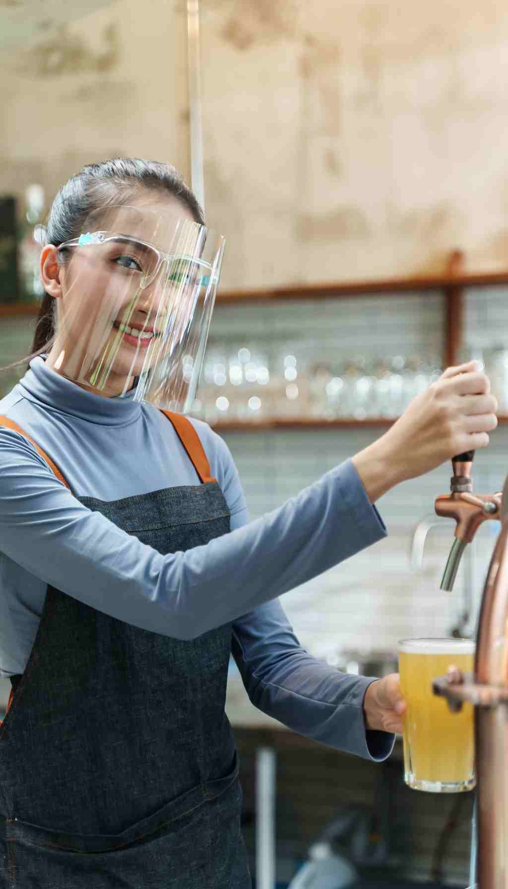 Bartender Pouring Draft Beer While Wearing a Face Shield A bartender wearing a protective face shield pours a glass of draft beer from a tap in a bar setting, demonstrating hygiene and safety procedures.