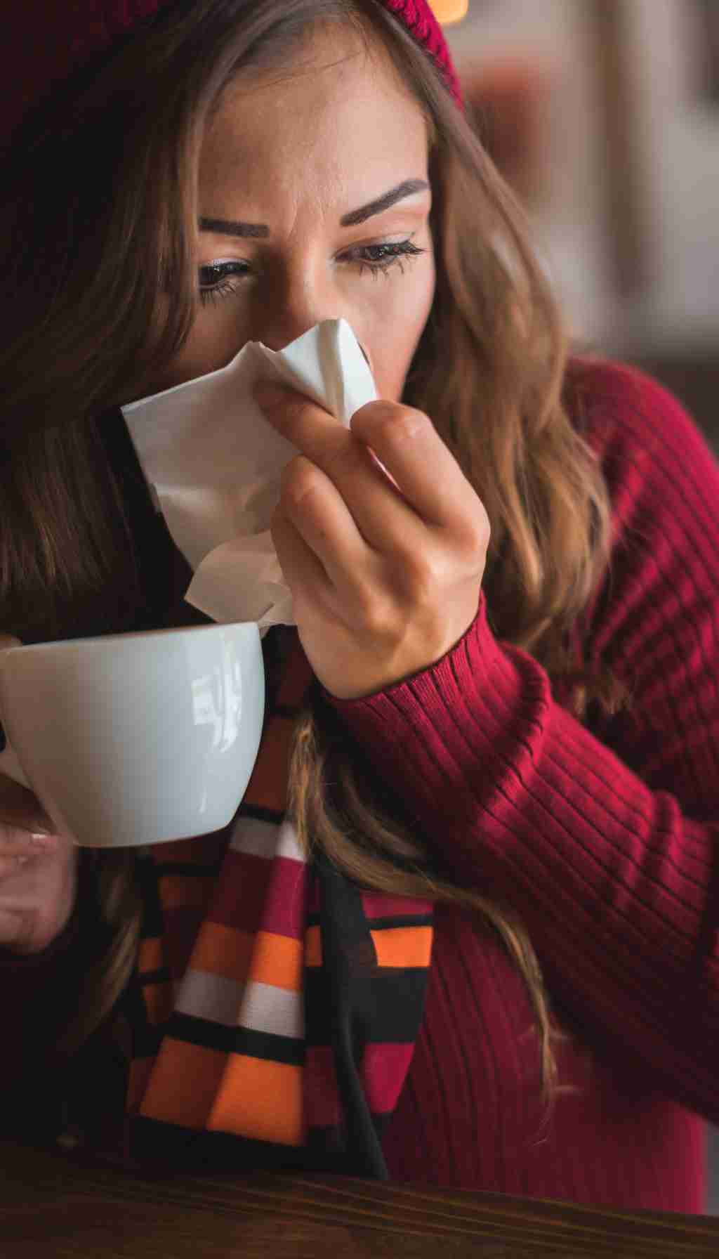 Woman With Cold Symptoms Holding a Hot Drink Indoors A woman in a red sweater holds a cup of hot tea while using a tissue to wipe her nose, showing visible cold or flu symptoms.
