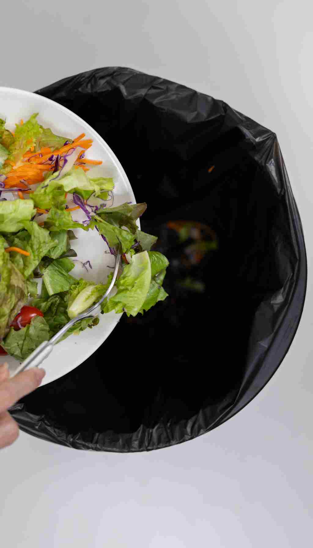 Discarded Salad Being Thrown Away in a Commercial Kitchen Bin A person scraping leftover salad from a plate into a black-lined bin, illustrating food waste.