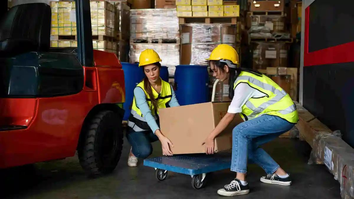 Two warehouse workers using team lifting techniques to move a heavy box safely onto a trolley in an industrial environment.