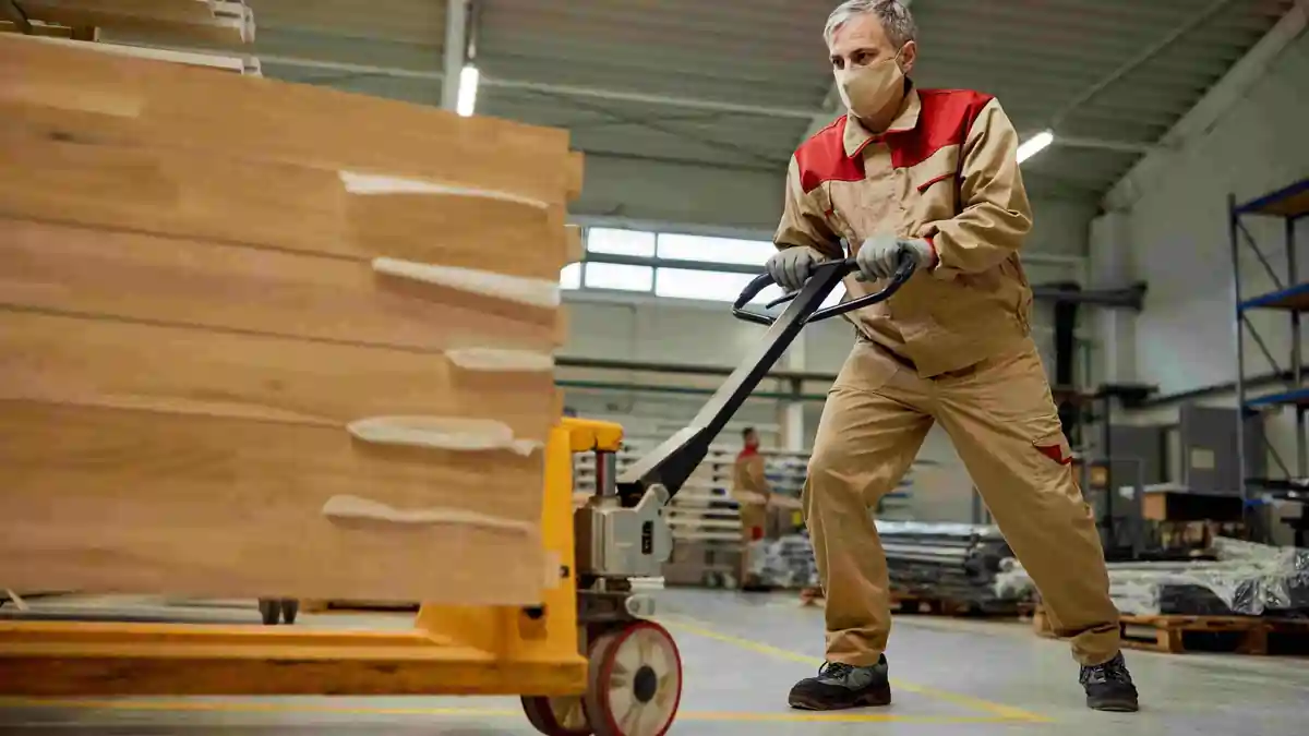 Worker using a pallet jack to move a heavy load of timber in a warehouse, demonstrating safe mechanical handling to reduce manual lifting risks.