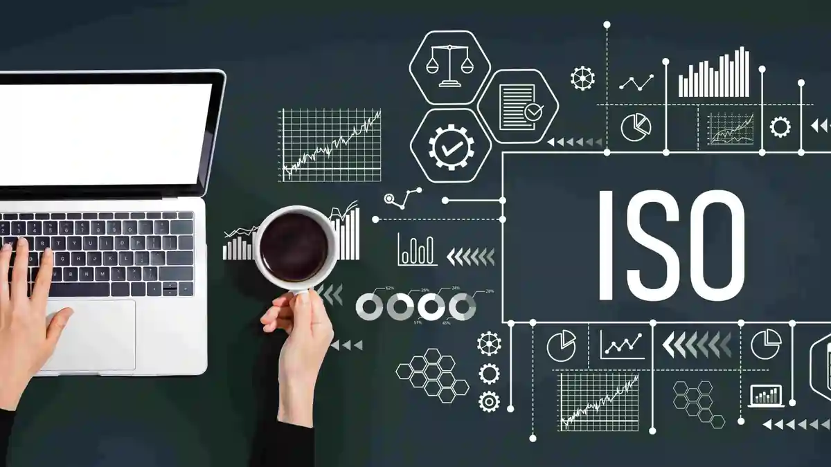ISO Standards and Digital Quality Management Illustration Hands typing on a laptop next to a cup of coffee, surrounded by digital icons, charts, and compliance graphics, with the word “ISO” displayed prominently on a dark background.