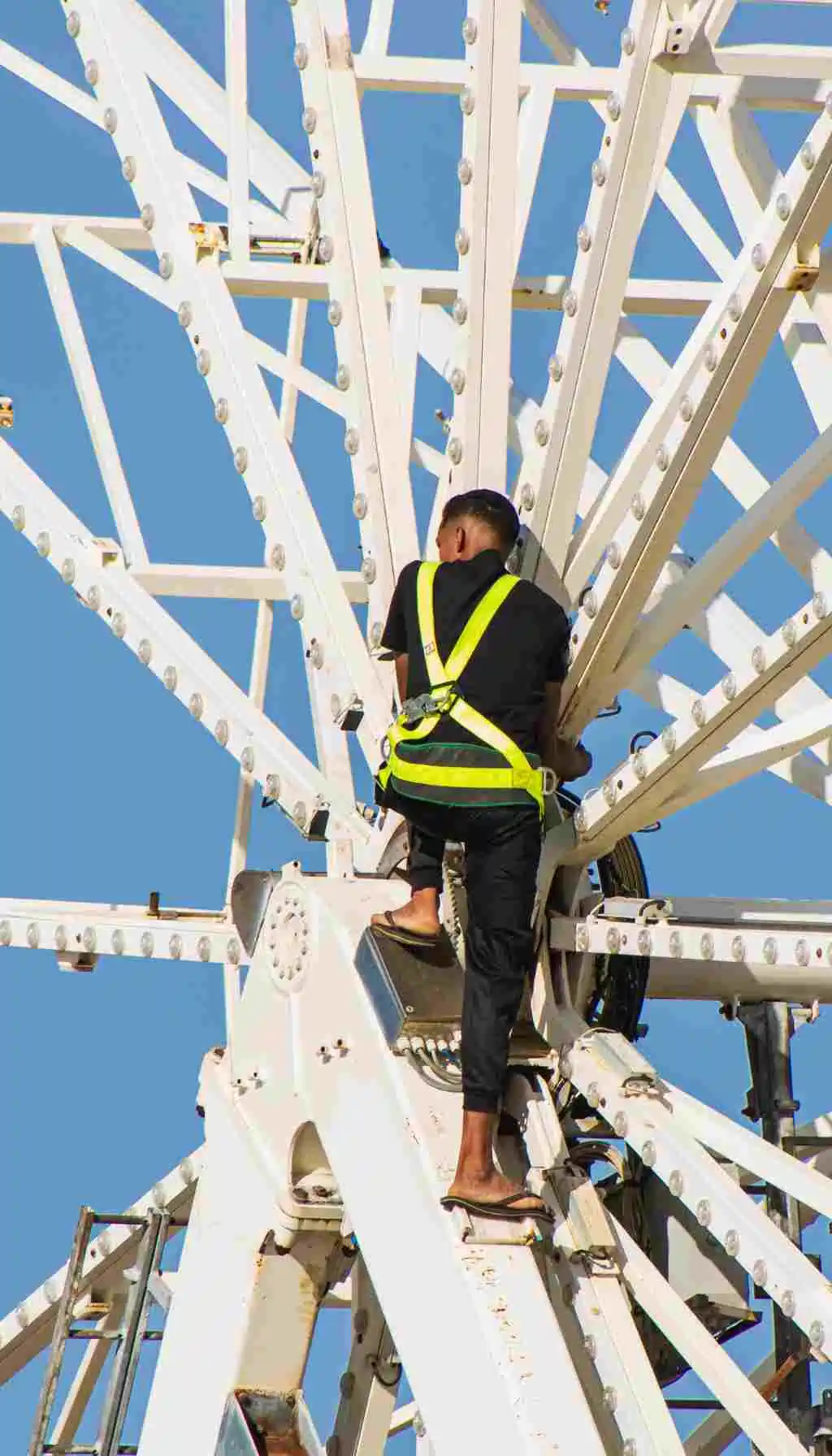 Worker on a high metal structure wearing a safety harness while performing maintenance at height under clear sky conditions.