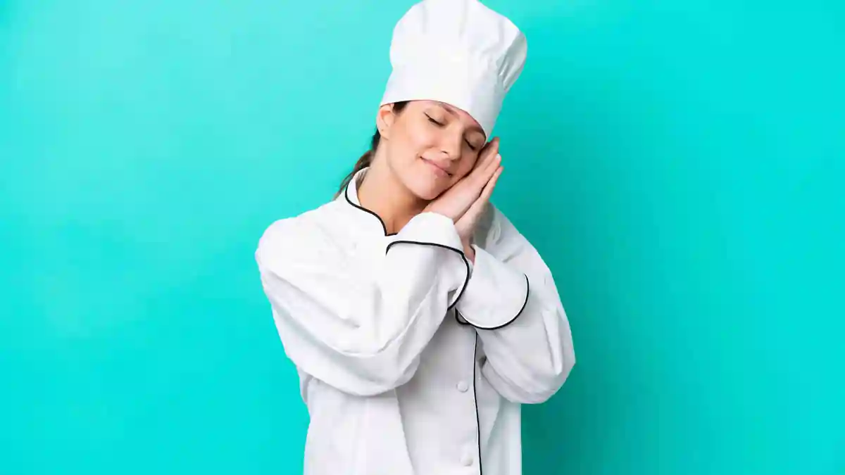 Chef in a white uniform and hat miming sleep with hands resting against face on a turquoise background