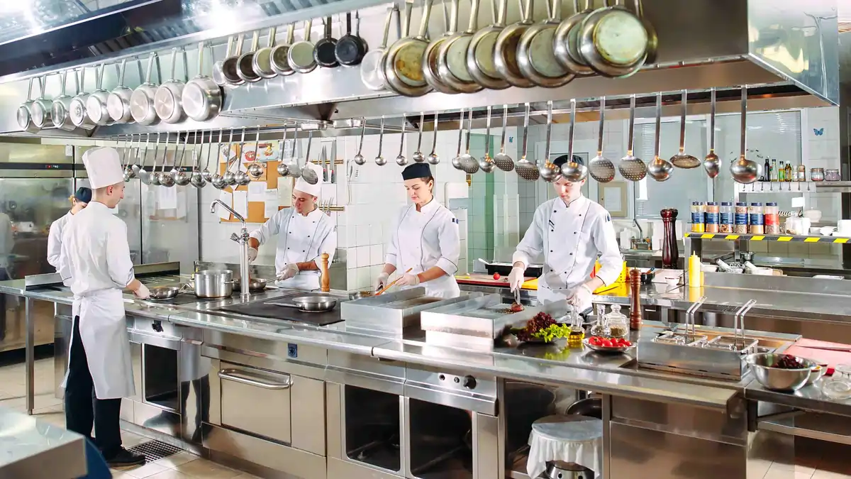 Commercial kitchen with chefs working at stainless steel workstations beneath hanging pans and utensils during food preparation