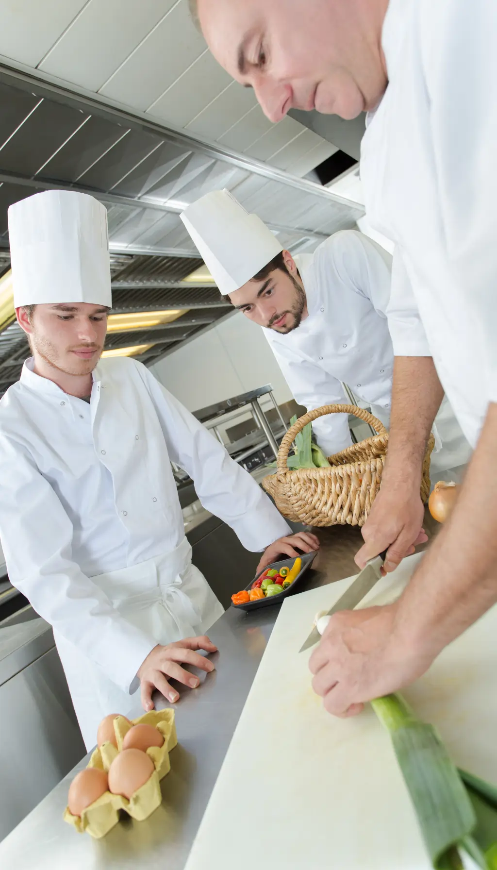 Head chef supervising junior chefs preparing fresh ingredients on a cutting board in a commercial kitchen.